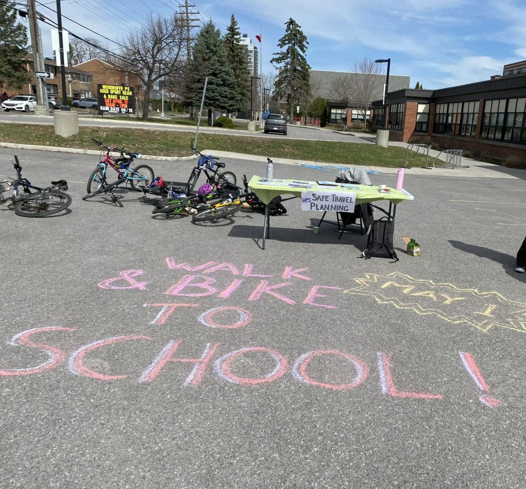 A picture of a school yard. In the forground is a messaged written in chalk "walk or bike to school". Kids bikes are scattered in the mid ground.